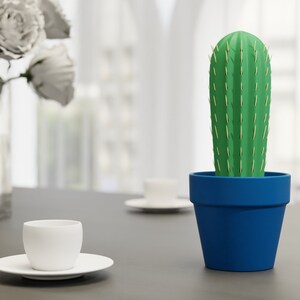 3D-gedruckter Kaktus Zahnstocherhalter in Topffarbe Marineblau auf einem Tisch neben einer weißen Tasse, imitiert mit herauskommenden Zahnstochern eine Kaktuspflanze.