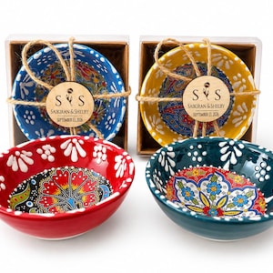 May include: Four small ceramic bowls featuring intricate floral patterns in red, blue, yellow, and teal. Two bowls are packaged in clear boxes with twine and wooden tags reading S S, SAIKIRAN & SHELBY, and SEPTEMBER 13, 2026. Two bowls sit in front.