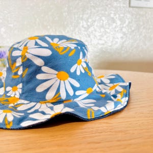 May include: A blue bucket hat with a white daisy pattern and yellow centers. The hat has a dark blue trim around the brim.