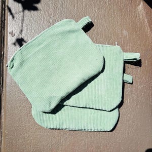 May include: Two sage green corduroy pot holders with a loop for hanging. The pot holders are rectangular with rounded corners and a ribbed texture. They are stacked on top of each other.