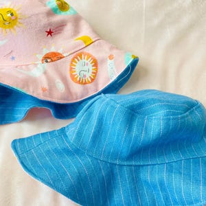 May include: Two reversible bucket hats. One hat features a pink fabric with a celestial print of suns, stars, and clouds. The other hat is blue with white pinstripes. Both hats have a blue brim.