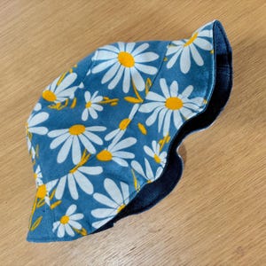 May include: A blue bucket hat with a white daisy floral pattern and a dark blue brim. The hat features a reversible design, showcasing a contrasting dark blue fabric on the inside. The daisies have yellow centers and yellow stems.