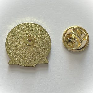 May include: The reverse of a gold-coloured pin with a round design. The pin has a post and clutch on the back.