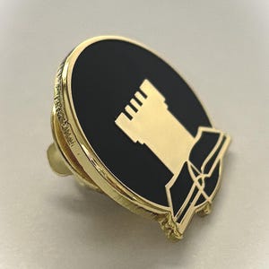 May include: Gold-coloured pin with a black enamel background. The pin features a white castle-like structure with a book-like shape below it.