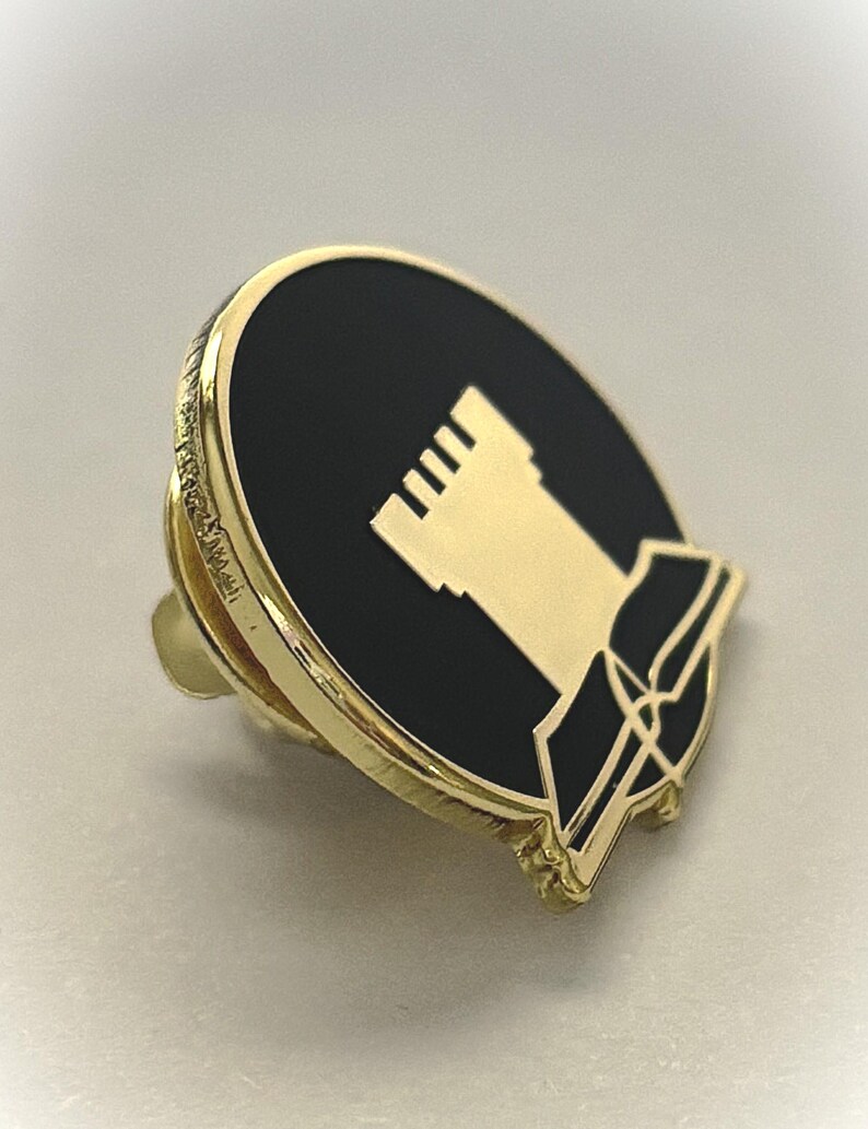 May include: Gold-coloured pin with a black enamel background. The pin features a white castle-like structure with a book-like shape below it.