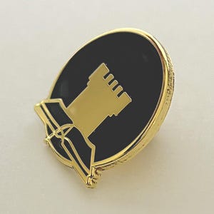 May include: Gold-coloured pin with a black enamel circle featuring a gold silhouette of a tower. The pin has a scroll-like design at the bottom.
