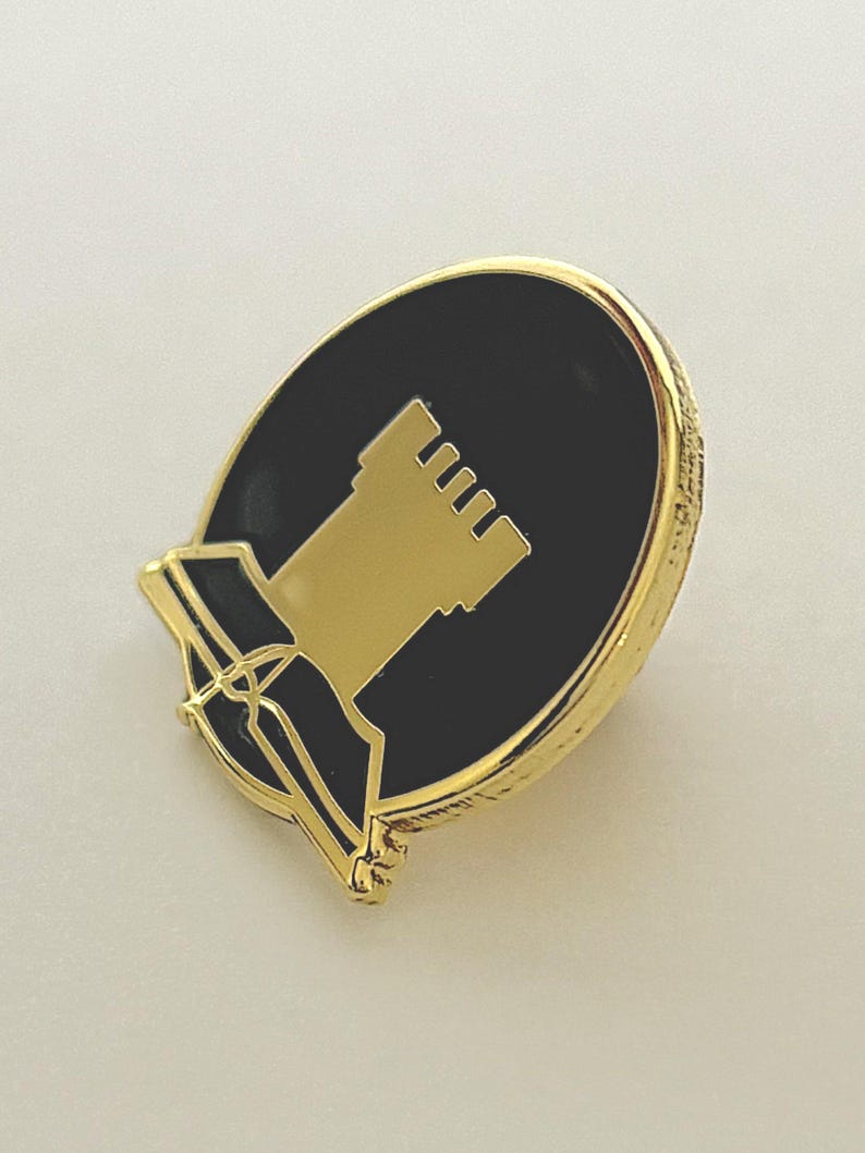 May include: Gold-coloured pin with a black enamel circle featuring a gold silhouette of a tower. The pin has a scroll-like design at the bottom.