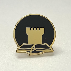 May include: Gold-coloured enamel pin featuring a black circle with a gold tower and a gold open book underneath.