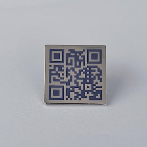 May include: Square metal pin with a QR code design. The pin has a silver border and a blue QR code pattern. The pin is set against a white background. Suitable for attaching to clothing or accessories.