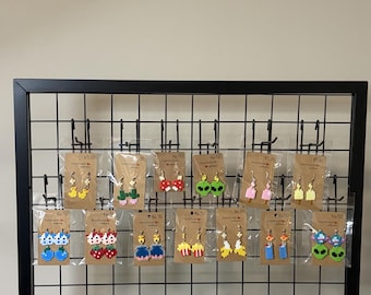 May include: A collection of pixel art style earrings made from small plastic beads, displayed on brown cardboard backing cards. The designs include various shapes like fruits, aliens, and objects, all hanging from metal hooks on a black wire grid.