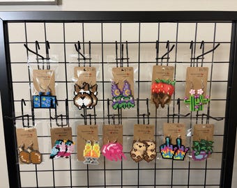 Op de afbeelding: Een collectie van elf paar handgemaakte pixel art oorbellen gemaakt van plastic kralen, gepresenteerd op bruine kartonnen kaartjes die aan een zwart metalen rooster hangen. De ontwerpen omvatten een sterrennacht, corgi, aardbei, cactus en meer.