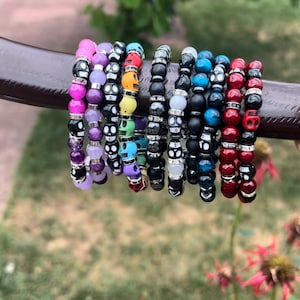 May include: A set of colorful skull bead bracelets with different gemstone accents. The bracelets are stacked on a brown surface.