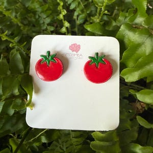 Peut inclure: Boucles d'oreilles rouges en forme de tomate avec des tiges vertes, présentées sur une carte blanche. Les boucles d'oreilles sont posées sur un fond de feuillage vert. Le texte "COSMEA ART STUDIO" est visible.