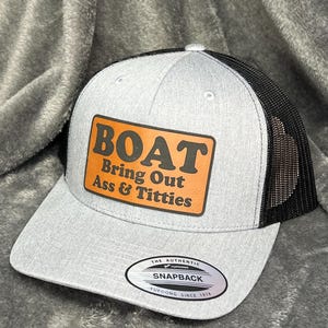 May include: A gray snapback hat with a black mesh back. The front features a brown rectangular patch with the words "BOAT Bring Out Ass & Titties" in bold orange letters. The hat has a curved brim and a snap closure.