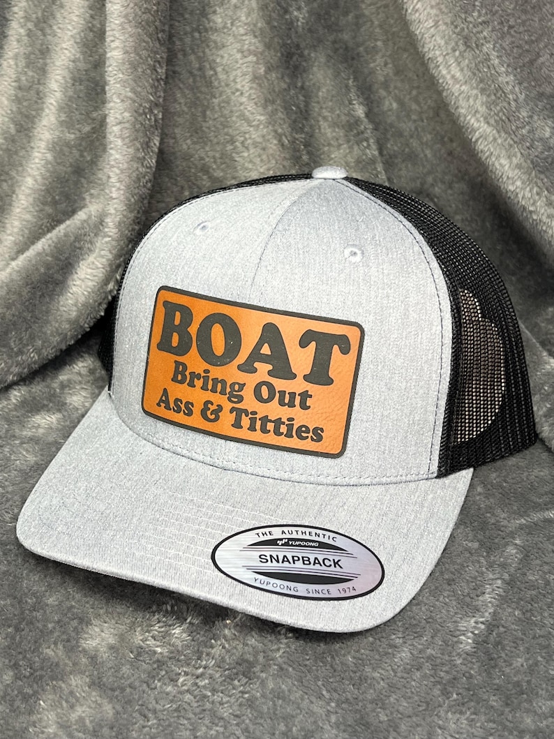 Może przedstawiać: Szara czapka typu snapback z czarną siatką z tyłu. Z przodu brązowa prostokątna naszywka z pomarańczowym napisem "BOAT Bring Out Ass & Titties". Czapka ma zakrzywiony daszek i zapięcie na zatrzask.