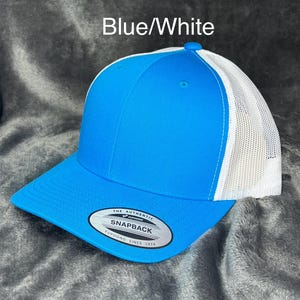 Może przedstawiać: Niebiesko-biała czapka typu snapback z zakrzywionym daszkiem. Przedni panel i daszek są w kolorze jasnoniebieskim, a tył z białej siatki. Czapka ma napis "Blue/White" i etykietę "Snapback".
