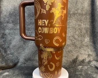 May include: A brown travel mug with a gold design featuring horses, horseshoes, and the text "Hey, Cowboy".