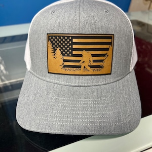 May include: A gray and white trucker hat with a brown leather patch featuring a silhouette of Bigfoot walking in front of a gold American flag.