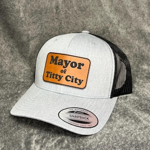 May include: A light gray trucker hat with a black mesh back and a curved brim. The hat features a brown leather patch with the words "Mayor of Titty City" in black lettering. The hat has a snapback closure.