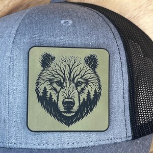 May include: A gray trucker hat with a black mesh back. The hat features a square patch with a detailed black bear face design on a green background. The bear's fur is depicted with tree-like patterns.