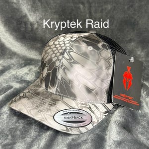 Może przedstawiać: Czapka z daszkiem Kryptek Raid z kamuflażem w odcieniach szarości i czerni. Czapka ma czarną siatkę z tyłu i zakrzywiony daszek. Widoczny jest napis "Kryptek Raid". Czapka ma metkę z napisem "Kryptek".