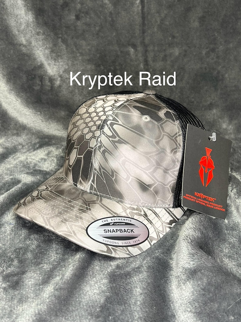Może przedstawiać: Czapka z daszkiem Kryptek Raid z kamuflażem w odcieniach szarości i czerni. Czapka ma czarną siatkę z tyłu i zakrzywiony daszek. Widoczny jest napis "Kryptek Raid". Czapka ma metkę z napisem "Kryptek".