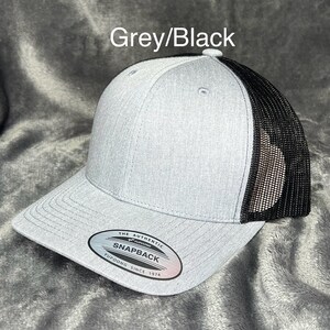 Może przedstawiać: Szara i czarna czapka z daszkiem Snapback. Prz&oacute;d czapki jest szary, z czarną siatką z tyłu. Czapka ma zakrzywiony daszek i zapięcie Snapback. Tekst "Grey/Black" znajduje się nad czapką.