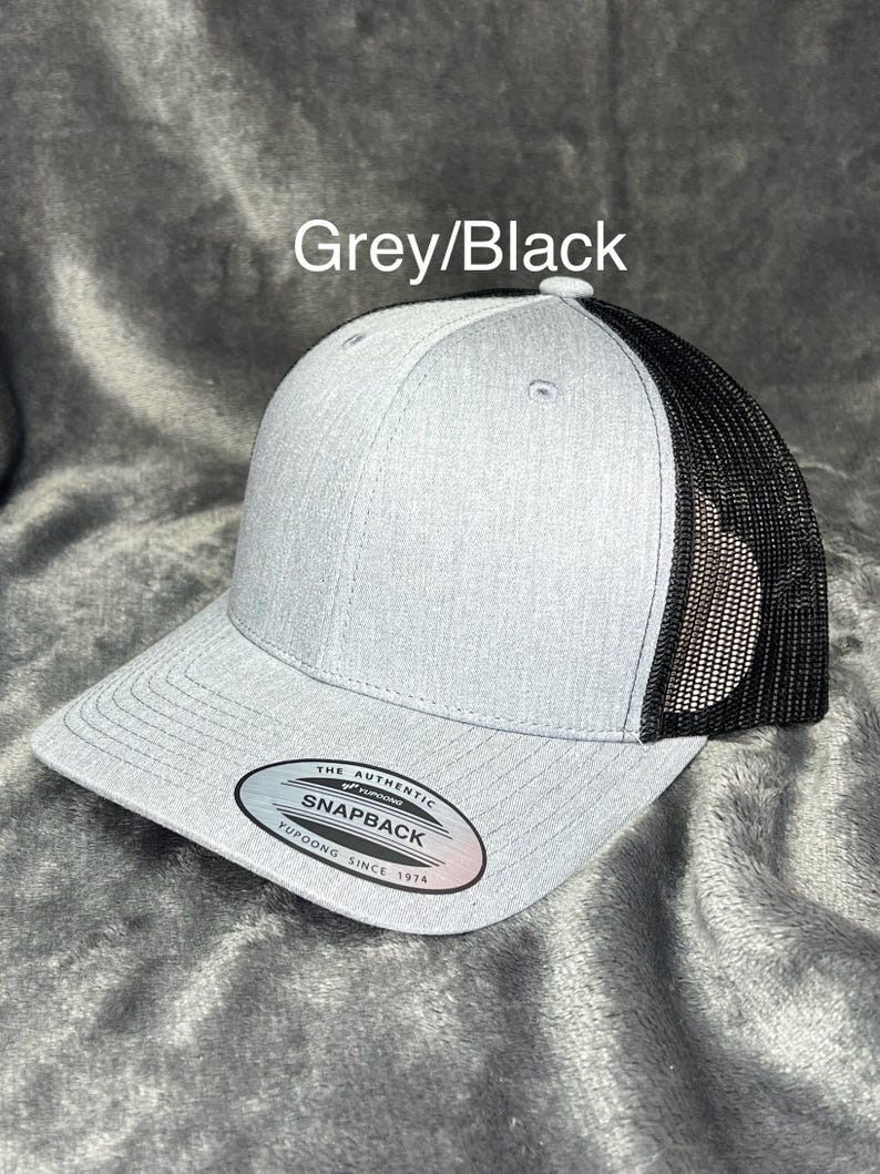 Może przedstawiać: Szara i czarna czapka z daszkiem Snapback. Prz&oacute;d czapki jest szary, z czarną siatką z tyłu. Czapka ma zakrzywiony daszek i zapięcie Snapback. Tekst "Grey/Black" znajduje się nad czapką.