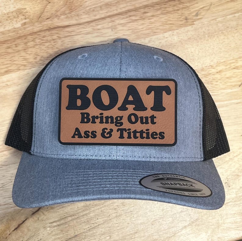 Może przedstawiać: Szara czapka typu trucker z czarną siatką z tyłu i brązową sk&oacute;rzaną naszywką. Na naszywce napis "BOAT" nad frazą "Bring Out Ass & Titties" w kolorze czarnym. Czapka ma zapięcie na zatrzask.