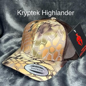 Może przedstawiać: Czapka z daszkiem Kryptek Highlander z kamuflażem w odcieniach brązu, beżu i zieleni. Czapka ma brązową siatkową tylną część i zakrzywiony daszek. Z przodu widnieje napis "Kryptek Highlander".
