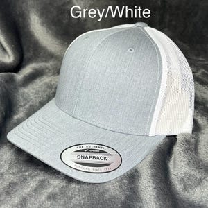 Może przedstawiać: Szara i biała czapka bejsbolowa Snapback. Przednie panele i daszek są szare, a tylna siatka biała. Czapka ma zakrzywiony daszek i zapięcie na zatrzask. Tekst "Grey/White" znajduje się nad czapką. Prz&oacute;d czapki ma czarno-białą owalną metkę z napisem "The Authentic Snapback Yupoong Since 1974".