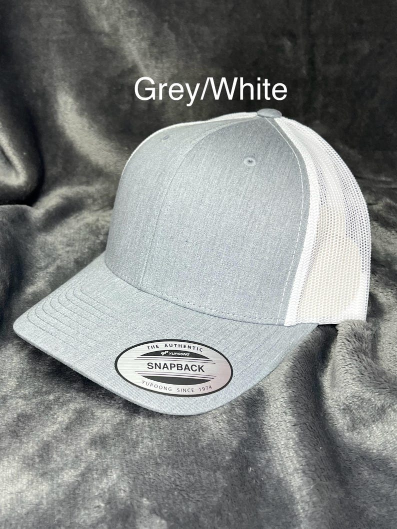 Może przedstawiać: Szara i biała czapka bejsbolowa Snapback. Przednie panele i daszek są szare, a tylna siatka biała. Czapka ma zakrzywiony daszek i zapięcie na zatrzask. Tekst "Grey/White" znajduje się nad czapką. Prz&oacute;d czapki ma czarno-białą owalną metkę z napisem "The Authentic Snapback Yupoong Since 1974".