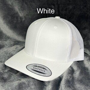 Może przedstawiać: Biała czapka bejsbolowa typu snapback z siatkowym tyłem. Czapka ma zakrzywiony daszek i strukturalny panel przedni. Na zdjęciu widnieje napis "White". Czapka ma etykietę "Snapback".