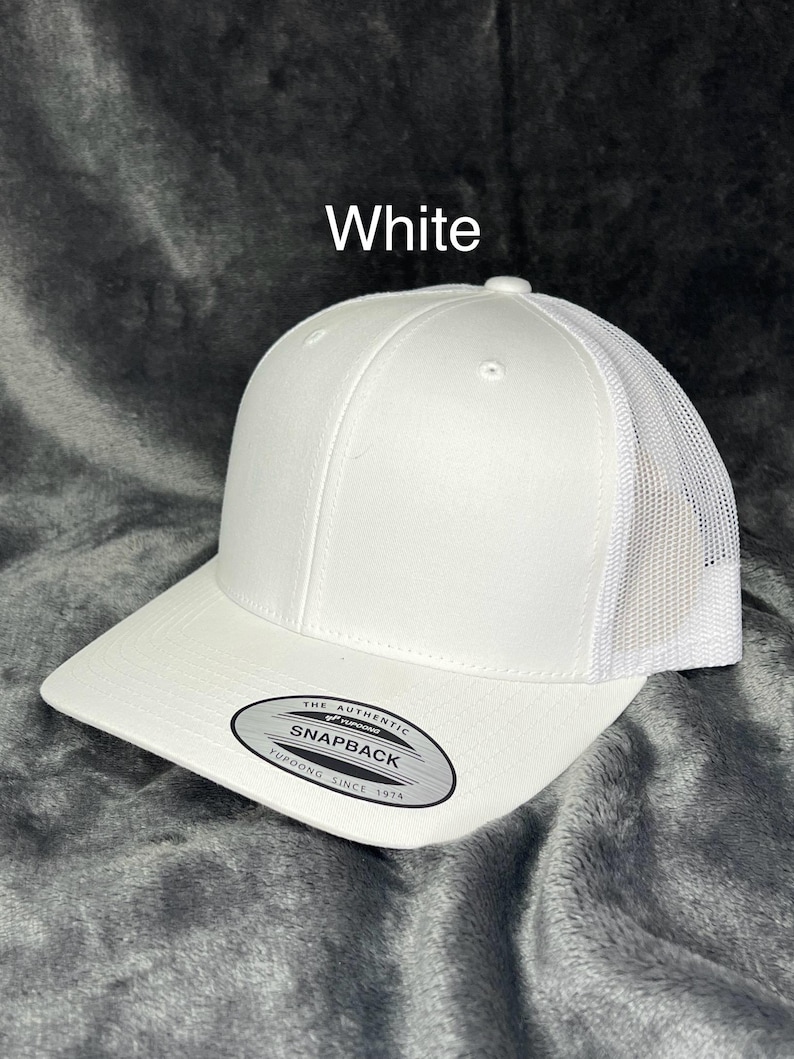 Może przedstawiać: Biała czapka bejsbolowa typu snapback z siatkowym tyłem. Czapka ma zakrzywiony daszek i strukturalny panel przedni. Na zdjęciu widnieje napis "White". Czapka ma etykietę "Snapback".