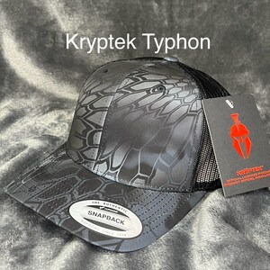 Może przedstawiać: Czarna i szara czapka z daszkiem Kryptek Typhon Snapback z siatkowym tyłem. Czapka ma unikalny geometryczny wz&oacute;r i napis "Kryptek Typhon". Zawiera metkę z logo Kryptek.