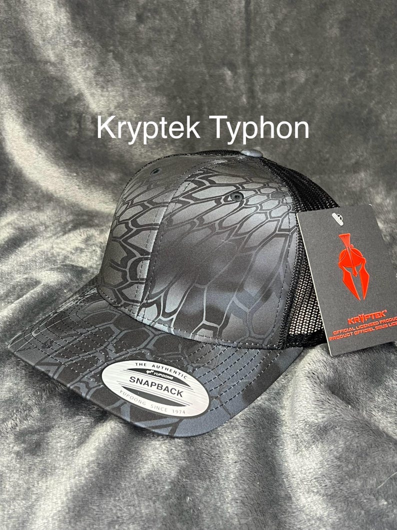 Może przedstawiać: Czarna i szara czapka z daszkiem Kryptek Typhon Snapback z siatkowym tyłem. Czapka ma unikalny geometryczny wz&oacute;r i napis "Kryptek Typhon". Zawiera metkę z logo Kryptek.