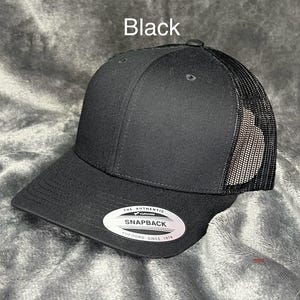 Może przedstawiać: Czarna czapka typu trucker z zakrzywionym daszkiem i siatkowym tyłem. Czapka ma napis "Black" i białą owalną etykietę z napisem "THE AUTHENTIC SNAPBACK YUPOONG SINCE 1974".