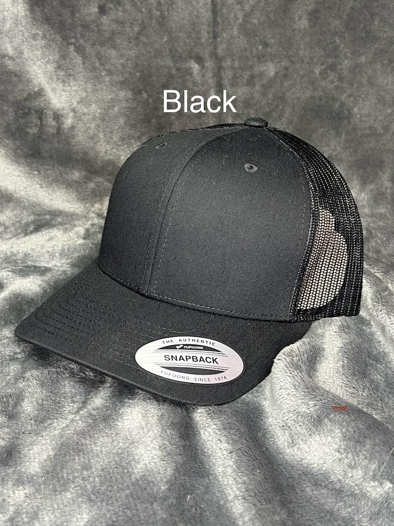Może przedstawiać: Czarna czapka typu trucker z zakrzywionym daszkiem i siatkowym tyłem. Czapka ma napis "Black" i białą owalną etykietę z napisem "THE AUTHENTIC SNAPBACK YUPOONG SINCE 1974".