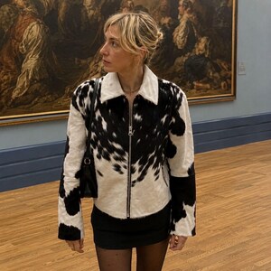 May include: A black and white cowhide-patterned jacket with a white collar and a silver zipper. The jacket is worn over a black skirt and tights. The background features a painting and a wooden floor.