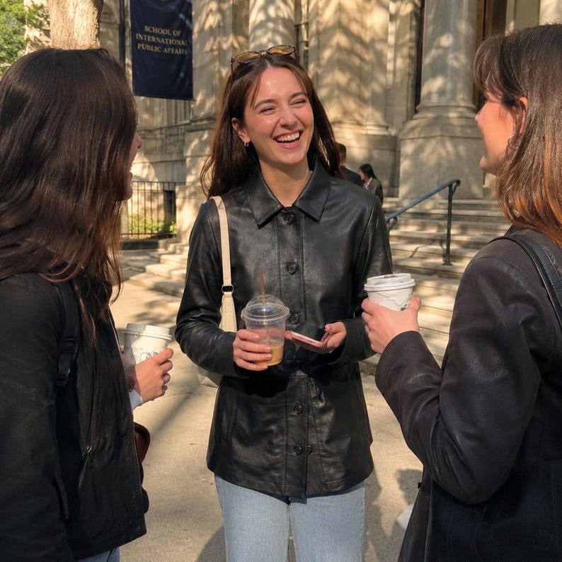 May include: Three people are standing outside a building, laughing and talking. One person is wearing a black leather jacket and light blue jeans, holding a clear plastic cup with a straw. The other two people are wearing black jackets and holding coffee cups.