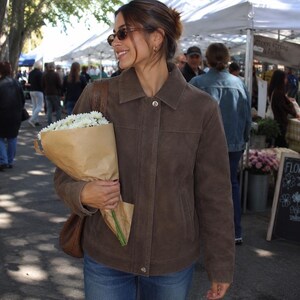 May include: A brown suede jacket with a collared neckline and button closures. The jacket is worn with blue jeans and a brown shoulder bag. A bouquet of white flowers wrapped in brown paper is held in the person's hand.
