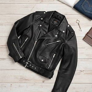 May include: Black leather motorcycle jacket with silver zippers, a belt, and snap details. A white t-shirt and blue jeans are visible in the background. The jacket is labeled "Handcrafted by KORDOVAN Leather Genuine Leather."