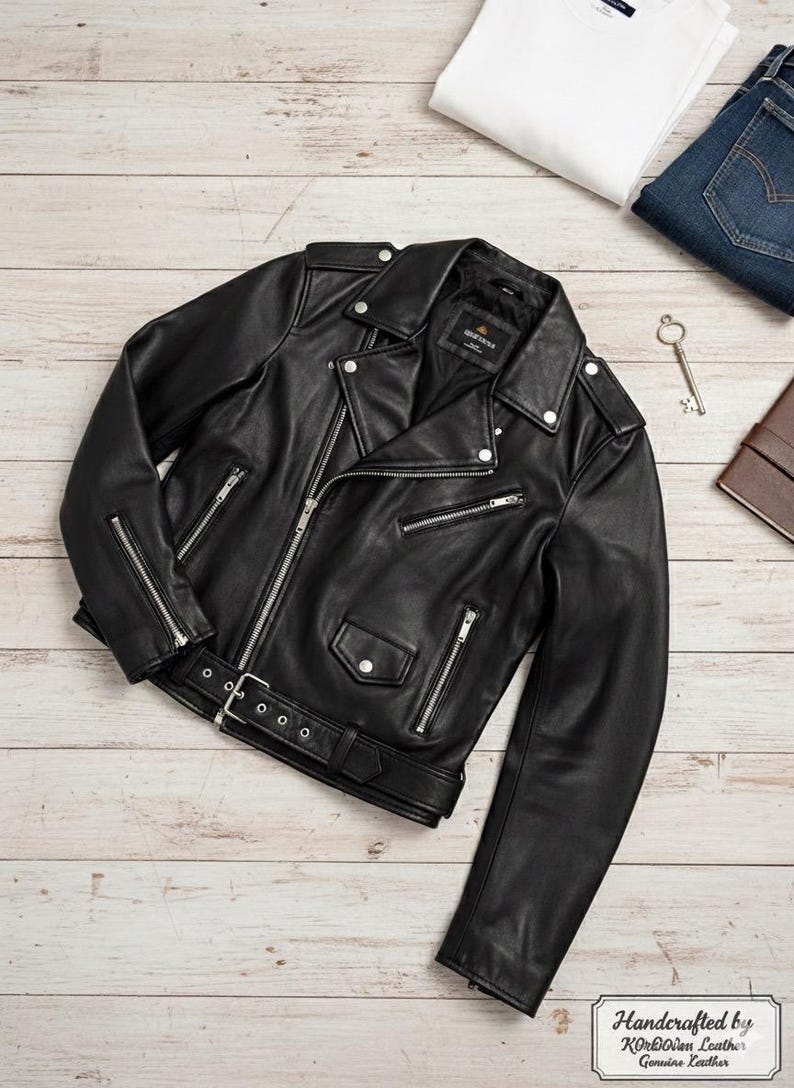 May include: Black leather motorcycle jacket with silver zippers, a belt, and snap details. A white t-shirt and blue jeans are visible in the background. The jacket is labeled "Handcrafted by KORDOVAN Leather Genuine Leather."