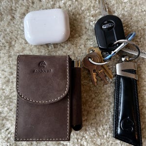 May include: A selection of everyday carry items. Includes a brown leather cardholder branded "Kordovan", a white wireless earbud case, a set of keys with a Ford key fob, and a black leather key holder.