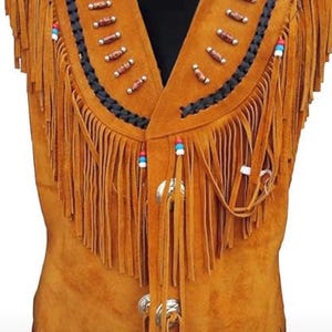 May include: A tan suede vest with a V-neck design. The vest features long fringe along the bottom and sides, with decorative beading and a black braided trim along the collar. Silver-colored metal clasps secure the front.
