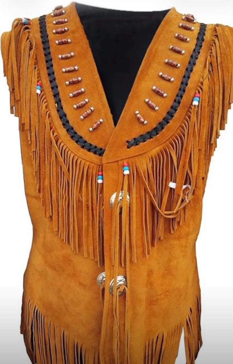 May include: A tan suede vest with a V-neck design. The vest features long fringe along the bottom and sides, with decorative beading and a black braided trim along the collar. Silver-colored metal clasps secure the front.