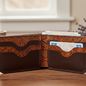 May include: Open brown leather wallet with snake-skin patterned accents. The wallet is open, revealing card slots and a section for cash. A credit card is visible in one of the slots. The wallet is on a wooden surface.