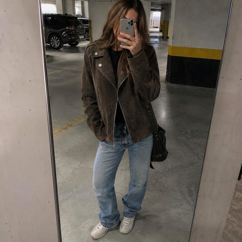 May include: A brown suede biker jacket with a silver zipper and snap details. The jacket is paired with light blue jeans and white sneakers. A black shoulder bag completes the outfit. The image is taken in a parking garage.