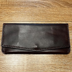 May include: A dark brown leather wallet with a flap closure. The rectangular wallet is hand-stitched with a contrasting thread. The leather has a smooth, slightly glossy finish.