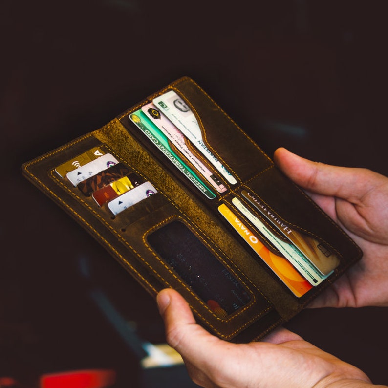 May include: Open, brown leather wallet filled with various credit cards, identification cards, and currency. The wallet is held open, showcasing multiple compartments and a clear ID window. The stitching is a contrasting yellow.